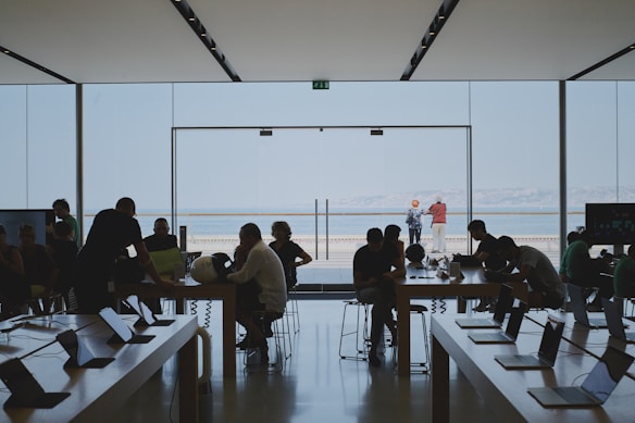 People are sitting and working at tables with laptops inside a store or office space with a large glass window overlooking an outdoor area. A few individuals are standing and interacting near the entrance, while two people are seen outside looking at the view.