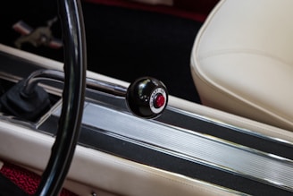 A close-up view of a vintage car interior featuring a manual gear shifter with a round knob labeled 'go baby go' in red. The seat is covered in white upholstery, and there are metallic details along the car's console.