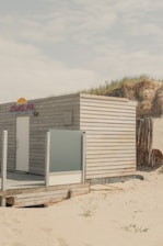 A small wooden beach hut with light-colored panels is situated on a sandy beach. The structure has a frosted glass panel on its porch and a sign with the text 'STRANDLOPER'. In the background, sand dunes topped with grass can be seen under a cloudy sky.