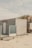 A small wooden beach hut with light-colored panels is situated on a sandy beach. The structure has a frosted glass panel on its porch and a sign with the text 'STRANDLOPER'. In the background, sand dunes topped with grass can be seen under a cloudy sky.