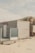 A small wooden beach hut with light-colored panels is situated on a sandy beach. The structure has a frosted glass panel on its porch and a sign with the text 'STRANDLOPER'. In the background, sand dunes topped with grass can be seen under a cloudy sky.
