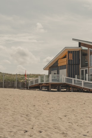 A wooden beach house elevated on stilts, with a modern design featuring large glass windows and navy blue paneling with contrasting yellow accents. A sandy beach stretches out in the foreground with a grassy dune in the background. A red flag is visible near the beach house, indicating a possible lifeguard post or signal.