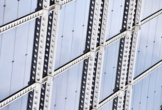 A close-up view of a modern architectural facade featuring repetitive geometric patterns. The structure comprises glass panels bordered by metallic beams with evenly spaced rivets, creating a grid-like appearance.