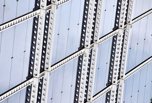 A close-up view of a modern architectural facade featuring repetitive geometric patterns. The structure comprises glass panels bordered by metallic beams with evenly spaced rivets, creating a grid-like appearance.