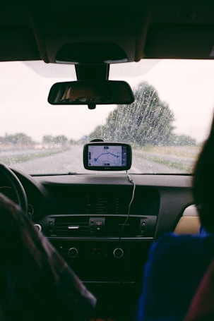 A view from inside a car, looking out through the windshield on a rainy day. A GPS device is mounted on the dashboard, displaying a map. Water droplets are visible on the glass, and there are two people in the front seats, one driver and one passenger.