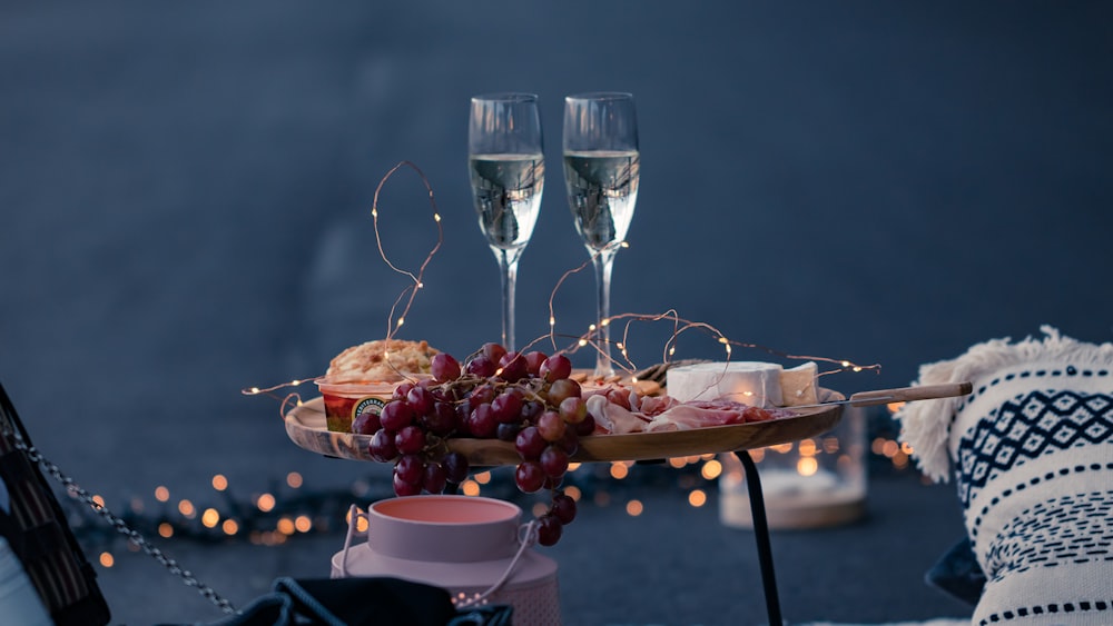 two glass of white wine near grapes fruit, string lights, and food on round wooden table beside white and black pillow