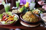 A variety of colorful dishes are presented on a wooden table, including a bowl of mixed salad with tomatoes, bell peppers, olives, and lettuce, alongside a plate of cooked legumes and grains topped with herbs. Green smoothies with lime slices are served in glasses, and a yellow flower centerpiece decorates the table.