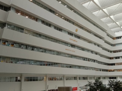 A large multi-story building interior with rows of windows and a sign indicating 'Ministry of Justice'. The architecture is modern with a white color scheme, and some greenery is visible at the bottom of the image.