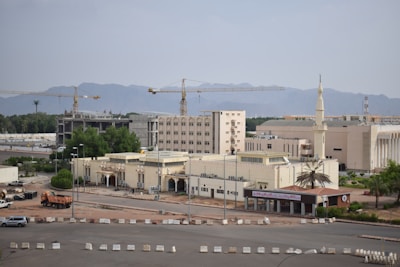 A construction site with several large yellow cranes towering over a complex of light-colored buildings. In the background, a range of mountains creates a natural backdrop. The area is surrounded by lush green trees, and there are vehicles parked near the buildings. A tall minaret is visible, indicating the presence of a mosque.