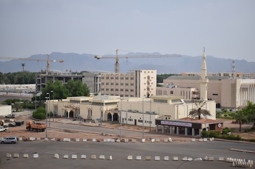 A construction site with several large yellow cranes towering over a complex of light-colored buildings. In the background, a range of mountains creates a natural backdrop. The area is surrounded by lush green trees, and there are vehicles parked near the buildings. A tall minaret is visible, indicating the presence of a mosque.