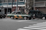 Two taxis are waiting at a crosswalk on a city street. One taxi has a green and orange color scheme, and the other is dark green with a logo referencing the Tokyo 2020 Olympics. The background includes buildings and a Panasonic sign.