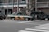 Two taxis are waiting at a crosswalk on a city street. One taxi has a green and orange color scheme, and the other is dark green with a logo referencing the Tokyo 2020 Olympics. The background includes buildings and a Panasonic sign.