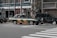 Two taxis are waiting at a crosswalk on a city street. One taxi has a green and orange color scheme, and the other is dark green with a logo referencing the Tokyo 2020 Olympics. The background includes buildings and a Panasonic sign.
