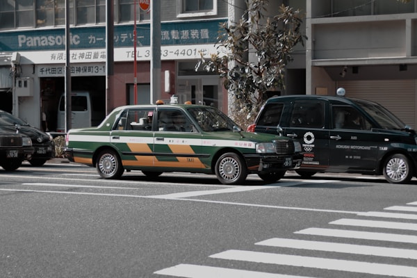 Two taxis are waiting at a crosswalk on a city street. One taxi has a green and orange color scheme, and the other is dark green with a logo referencing the Tokyo 2020 Olympics. The background includes buildings and a Panasonic sign.