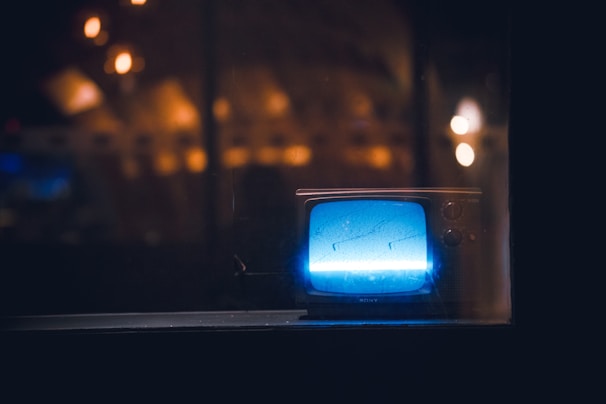A vintage television set is placed behind a glass pane, emitting a soft blue glow in a dimly lit environment. The background is filled with blurred, warm lights creating a cozy ambient effect.