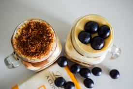 Two glass jars are filled with layered desserts, one topped with a crumbly brown topping, and the other with whole dark grapes. Additional grapes are scattered on the table along with a partially visible piece of packaging.