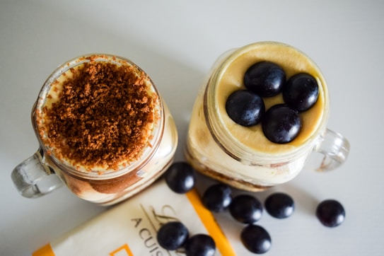 Two glass jars are filled with layered desserts, one topped with a crumbly brown topping, and the other with whole dark grapes. Additional grapes are scattered on the table along with a partially visible piece of packaging.