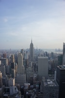 A vast skyline of a bustling city featuring numerous skyscrapers with the iconic structure of the Empire State Building prominently visible in the center. The cityscape extends towards a body of water under a partly cloudy sky.