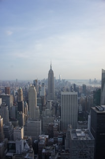 A vast skyline of a bustling city featuring numerous skyscrapers with the iconic structure of the Empire State Building prominently visible in the center. The cityscape extends towards a body of water under a partly cloudy sky.