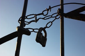 A close-up of a sturdy metal chain securing a lock, attached between metal poles, set against a clear blue sky.