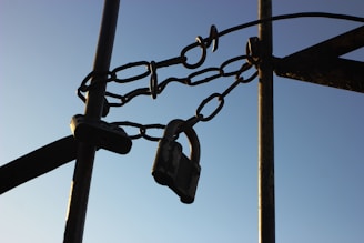 A close-up of a sturdy metal chain securing a lock, attached between metal poles, set against a clear blue sky.