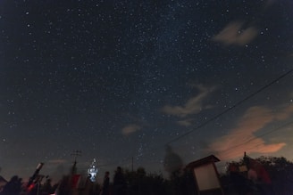 A dark sky filled with numerous stars, some cloud formations, and the faint outline of the Milky Way. Silhouettes of people and telescopes are noticeable in the foreground, suggesting an astronomy observation event. There are some visible wires crossing horizontally across the image.
