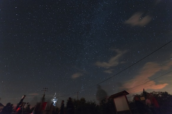 A dark sky filled with numerous stars, some cloud formations, and the faint outline of the Milky Way. Silhouettes of people and telescopes are noticeable in the foreground, suggesting an astronomy observation event. There are some visible wires crossing horizontally across the image.