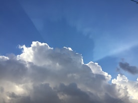 Large, fluffy cumulonimbus clouds with white tops and darker bases dominate the sky, contrasting against a backdrop of clear blue. Faint rays of sunlight can be seen breaking through, creating a serene and calming atmosphere. A subtle gradient from light to dark blue emphasizes the height and volume of the clouds.