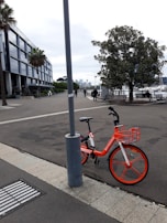 An urban scene featuring a vibrant orange rental bike parked against a lamppost. The area is a waterfront promenade with people walking and enjoying the outdoor space. A large tree and several benches are present, set against a backdrop of modern architecture and a hazy skyline.