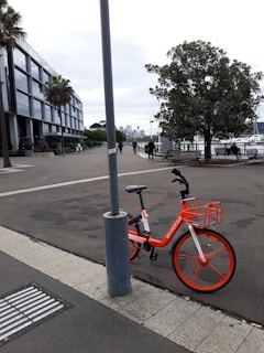 An urban scene featuring a vibrant orange rental bike parked against a lamppost. The area is a waterfront promenade with people walking and enjoying the outdoor space. A large tree and several benches are present, set against a backdrop of modern architecture and a hazy skyline.