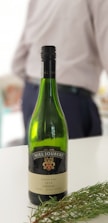 A bottle of Niel Joubert 2014 Shiraz wine is placed on a flat surface alongside some greenery. In the background, a person is partially visible, wearing a light-colored shirt and dark pants.