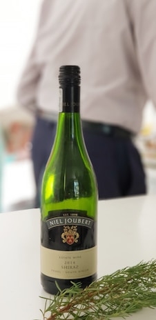 A bottle of Niel Joubert 2014 Shiraz wine is placed on a flat surface alongside some greenery. In the background, a person is partially visible, wearing a light-colored shirt and dark pants.