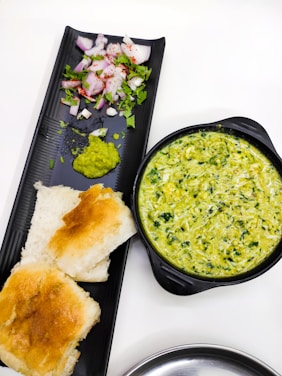 A black tray holds a portion of chopped onions sprinkled with red chili powder and garnished with coriander, along with a small amount of green chutney. Beside the tray, there is a black bowl filled with a creamy spinach and cheese dip. Two pieces of toasted bread are placed next to the dip on the tray.