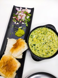 A black tray holds a portion of chopped onions sprinkled with red chili powder and garnished with coriander, along with a small amount of green chutney. Beside the tray, there is a black bowl filled with a creamy spinach and cheese dip. Two pieces of toasted bread are placed next to the dip on the tray.