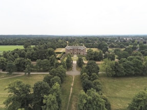 An aerial view of a large estate surrounded by dense greenery, featuring a grand building at the center. The area is filled with expansive lawns and a variety of trees, extending far into the horizon.