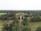 An aerial view of a large estate surrounded by dense greenery, featuring a grand building at the center. The area is filled with expansive lawns and a variety of trees, extending far into the horizon.
