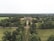 An aerial view of a large estate surrounded by dense greenery, featuring a grand building at the center. The area is filled with expansive lawns and a variety of trees, extending far into the horizon.