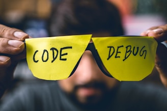 A person holds two bright yellow sticky notes shaped like glasses, with the word 'CODE' written on the left lens and 'DEBUG' on the right lens. The background is blurred, focusing attention on the makeshift glasses.