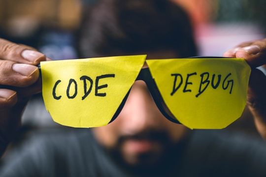 A person holds two bright yellow sticky notes shaped like glasses, with the word 'CODE' written on the left lens and 'DEBUG' on the right lens. The background is blurred, focusing attention on the makeshift glasses.