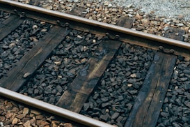 Weathered wooden railway sleepers are laid perpendicular to the steel rails, surrounded by an uneven bed of dark gravel and stones. The texture of the aged wood and the roughness of the stones contribute to an industrial and utilitarian aesthetic.