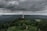 A telecommunications tower is prominently located on a hill surrounded by dense, lush green forest. The landscape extends into the distance with rolling hills under a dramatic, cloudy sky, creating a sense of vastness and isolation.