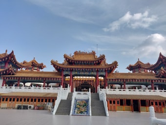 A traditional Chinese temple with ornate architectural features including intricate roof details and vibrant decorations. Stairs lead up to the entrance, flanked by decorative pillars and motifs. The setting is peaceful, under a blue sky with some clouds.