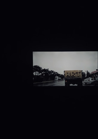 A road scene with a truck carrying stacked wooden crates driving along a wet street. Trees line the side of the road, and a few cars are parked nearby. The sky is overcast, contributing to a dim ambiance.