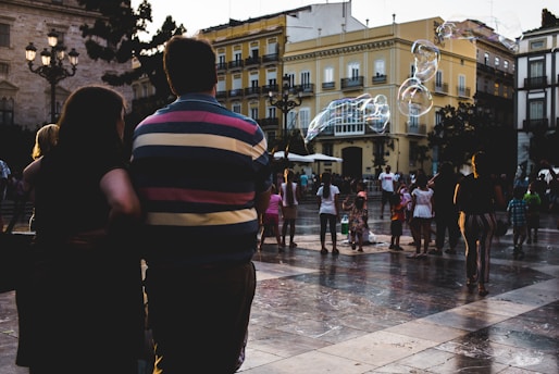 A group of people are gathered in a public square, possibly engaged in leisure activities. The scene includes large soap bubbles floating in the air. The background consists of historic buildings with ornate architecture, and the ground is made of stone or tile.