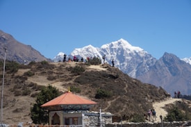 A group of hikers is walking along a trail on a hilly landscape with snow-capped mountains in the background. The foreground features a small structure with a decorative red roof.