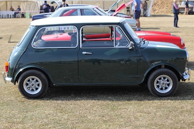 A classic dark green Mini Cooper is parked on grass. The car has a white roof and several colorful stickers on the rear side window. In the background, other vintage cars are visible alongside people standing near a tented area.