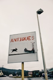 A sign with the word 'ANTIQUES' in large red letters, featuring a graphic of a dog and a scooter. The sign is mounted on a pole alongside a street light. The background shows a cloudy sky and a distant view of roads, vehicles, and surrounding buildings with another antique store further away.