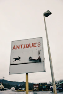 A sign with the word 'ANTIQUES' in large red letters, featuring a graphic of a dog and a scooter. The sign is mounted on a pole alongside a street light. The background shows a cloudy sky and a distant view of roads, vehicles, and surrounding buildings with another antique store further away.