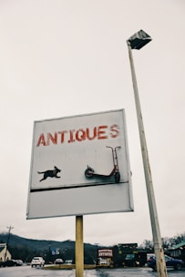 A sign with the word 'ANTIQUES' in large red letters, featuring a graphic of a dog and a scooter. The sign is mounted on a pole alongside a street light. The background shows a cloudy sky and a distant view of roads, vehicles, and surrounding buildings with another antique store further away.