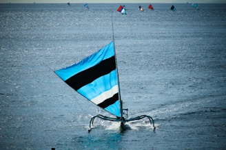 A sailboat with a large blue and black striped sail glides across a vast expanse of calm water. Several other sailboats with colorful sails are visible in the background, scattered across the sea. Sunlight reflects off the water, creating a shimmering effect.